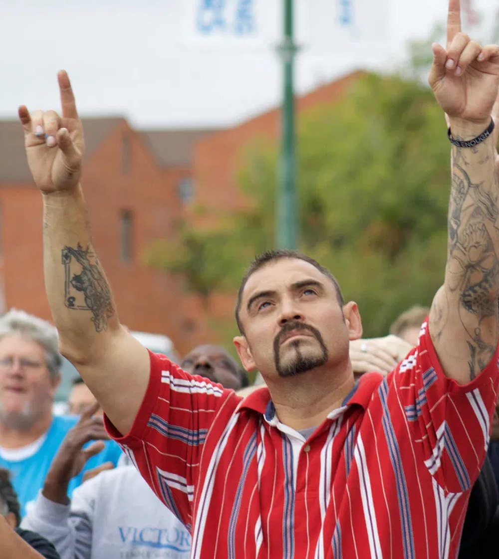 A man in a red striped shirt with both arms raised and tattoos down one forearm, eyes lifted, worshipping outdoors among a crowd in Victory Ministries shirts.